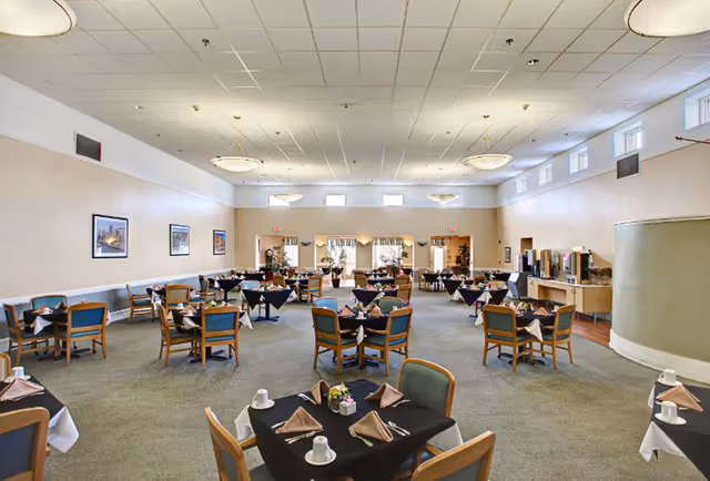 A spacious dining room with multiple tables covered in black tablecloths and neatly arranged with brown napkins, white cups, and silverware. The room has beige walls, several framed pictures, large windows near the ceiling, and ceiling lights. There is a buffet area on the right side of the room.