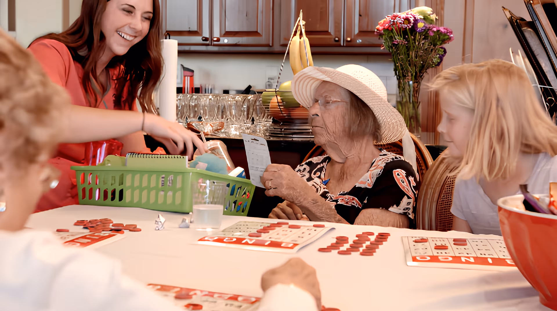 An elderly woman wearing a hat is sitting at a table playing bingo with children and a smiling caregiver in a kitchen setting. The table is covered with bingo cards and red markers, and there are flowers and kitchen cabinets in the background.