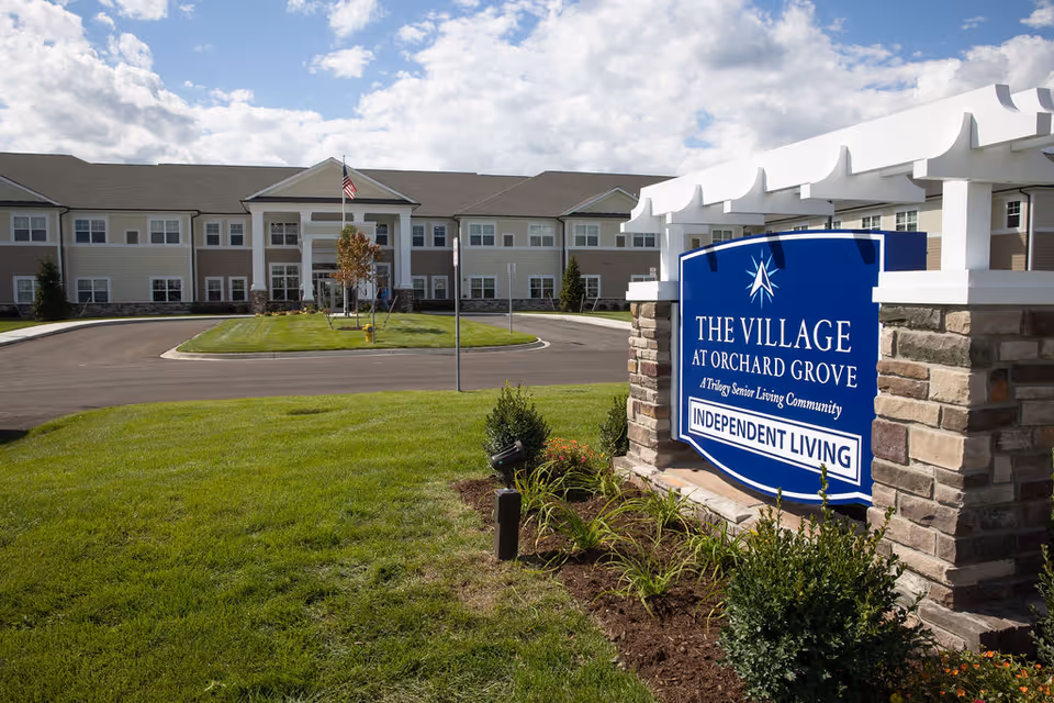 Exterior view of The Village at Orchard Grove senior living community building with a large blue sign in the foreground that reads 'The Village at Orchard Grove, A Trilogy Senior Living Community, Independent Living'. The building is two stories with multiple windows and an American flag flying in front. The sky is partly cloudy and the lawn is green and well-maintained.