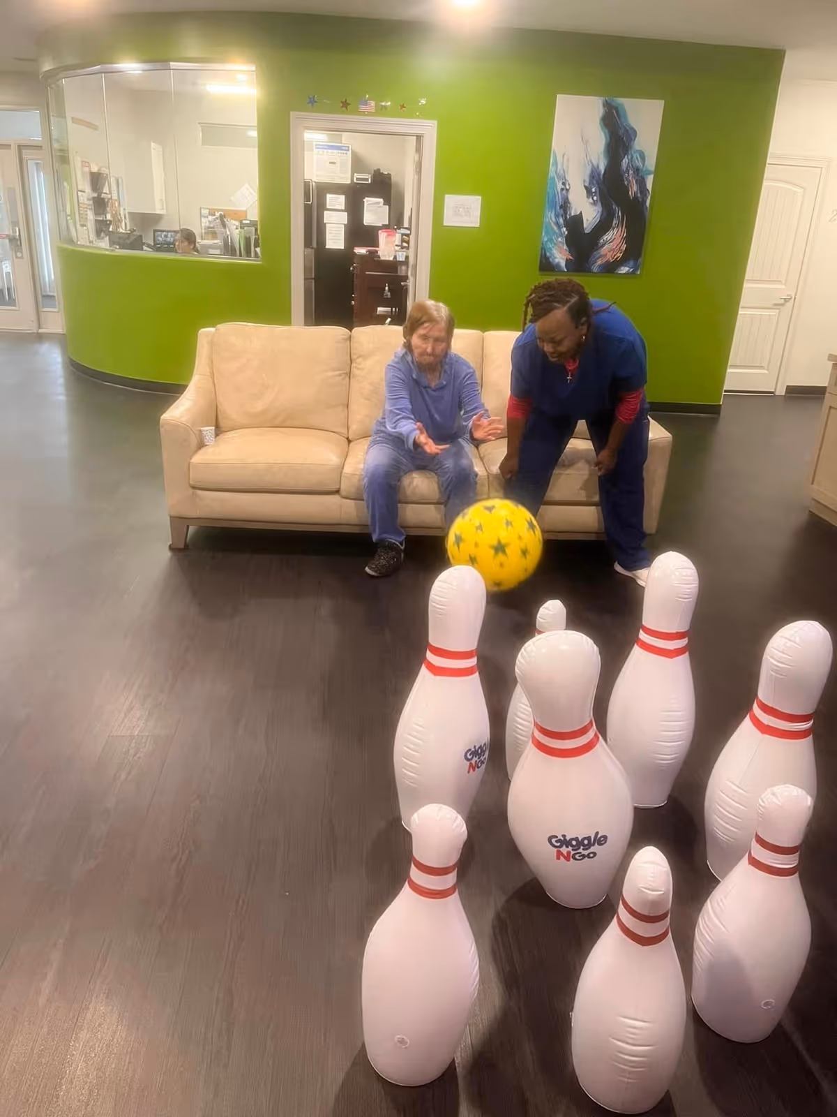 A resident and a caregiver play indoor bowling with a yellow ball and inflatable pins in a lounge area with a beige sofa and green wall.