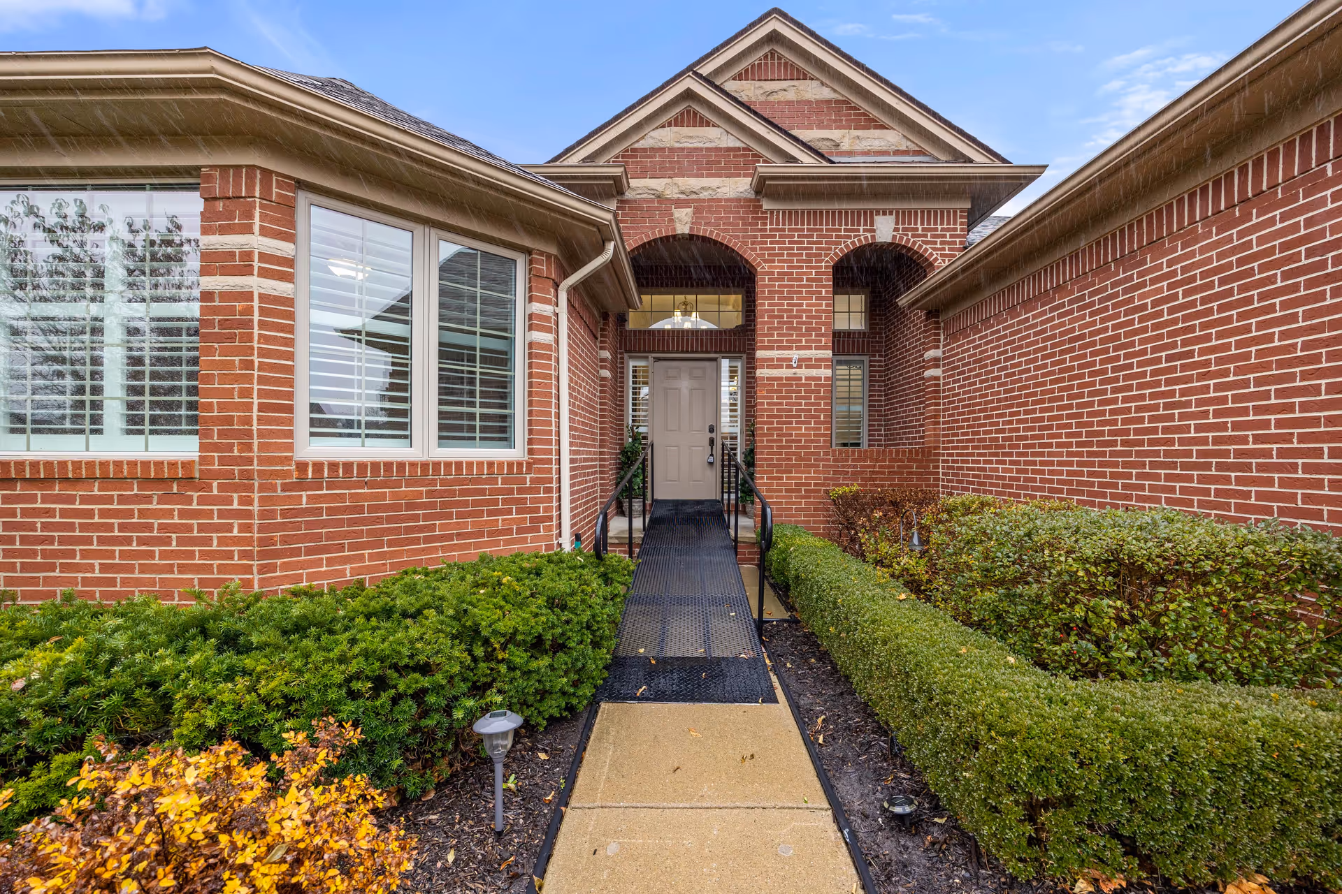 Front entrance of a brick building with a ramp leading up to a beige door. The entrance is framed by two brick arches and surrounded by neatly trimmed green bushes and plants. The sky is partly cloudy.