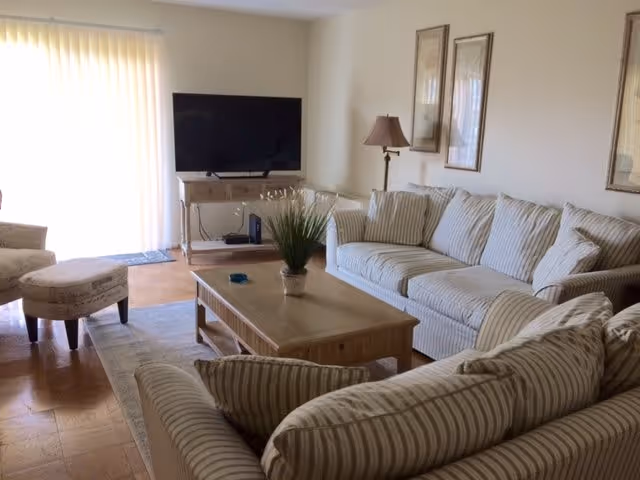 Bright living room with striped sofas, a wooden coffee table, TV on a stand, and a sliding glass door with vertical blinds.