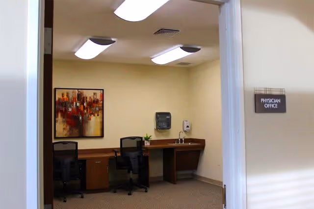 Interior view of a physician office with two black office chairs, a wooden desk, a small plant, a wall-mounted paper towel dispenser, and a hand sanitizer dispenser. There is a colorful abstract painting on the wall and fluorescent ceiling lights.