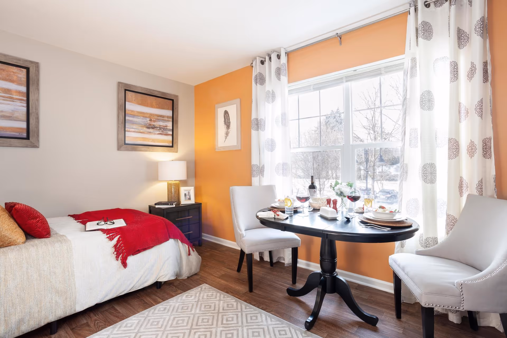 A cozy room with a single bed covered in beige and red bedding, a small black nightstand with a lamp and framed photo, and a round black dining table set for two with wine glasses and plates. The room has an orange accent wall, large window with white curtains featuring a circular pattern, and wooden flooring with a patterned rug.