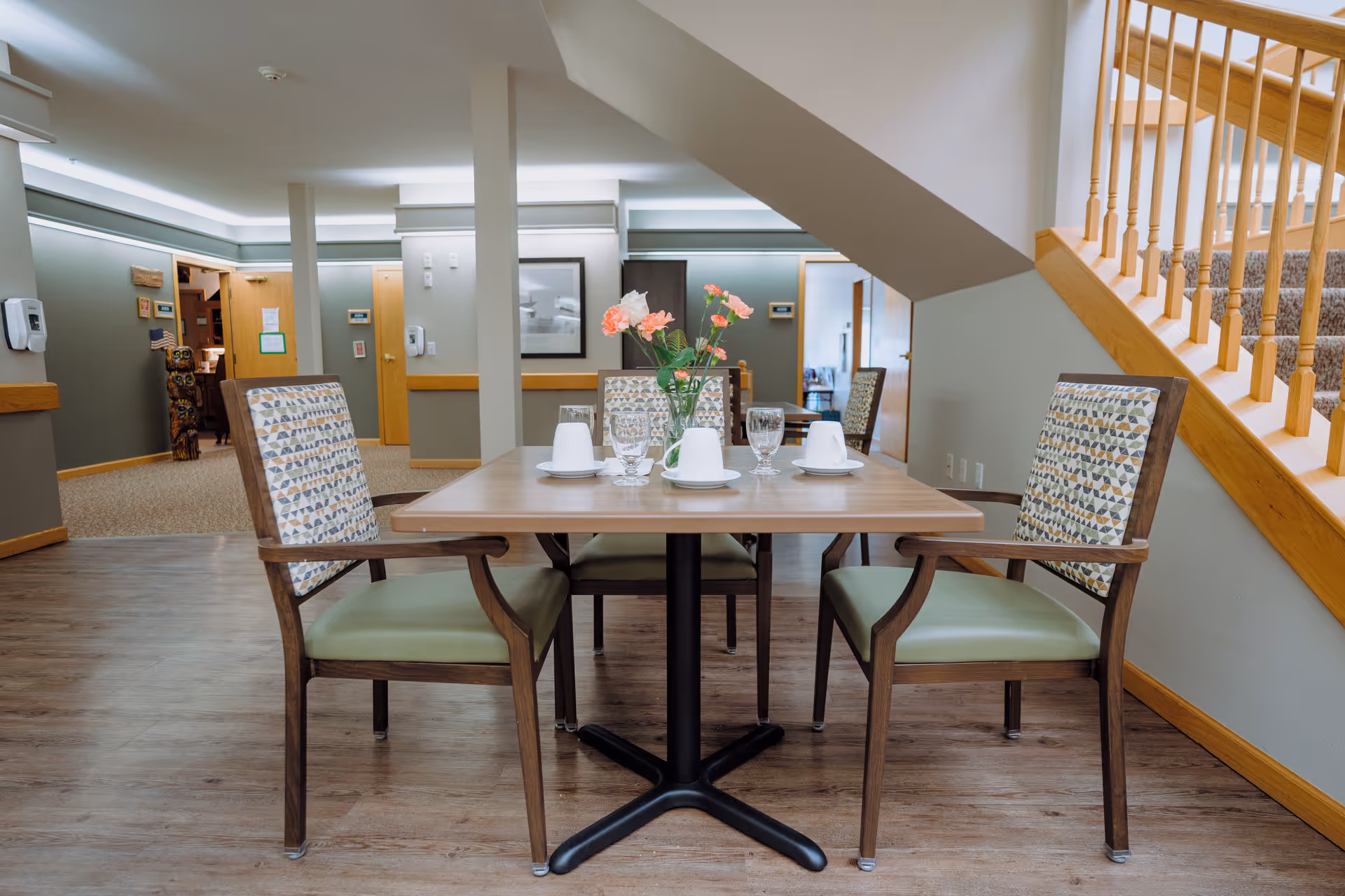 A dining area in a senior living facility with a wooden table set for four, featuring four chairs with patterned backs and green cushions. The table has a vase with pink flowers, upside-down cups on saucers, and water glasses. The room has wooden flooring, light-colored walls, and a staircase with wooden railings on the right side. The background shows doorways and a hallway with soft lighting.