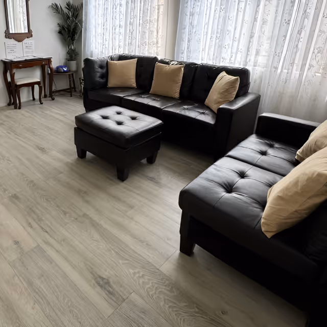 A bright living room with black leather sofas and an ottoman, beige throw pillows, light wood flooring and sheer curtained windows.