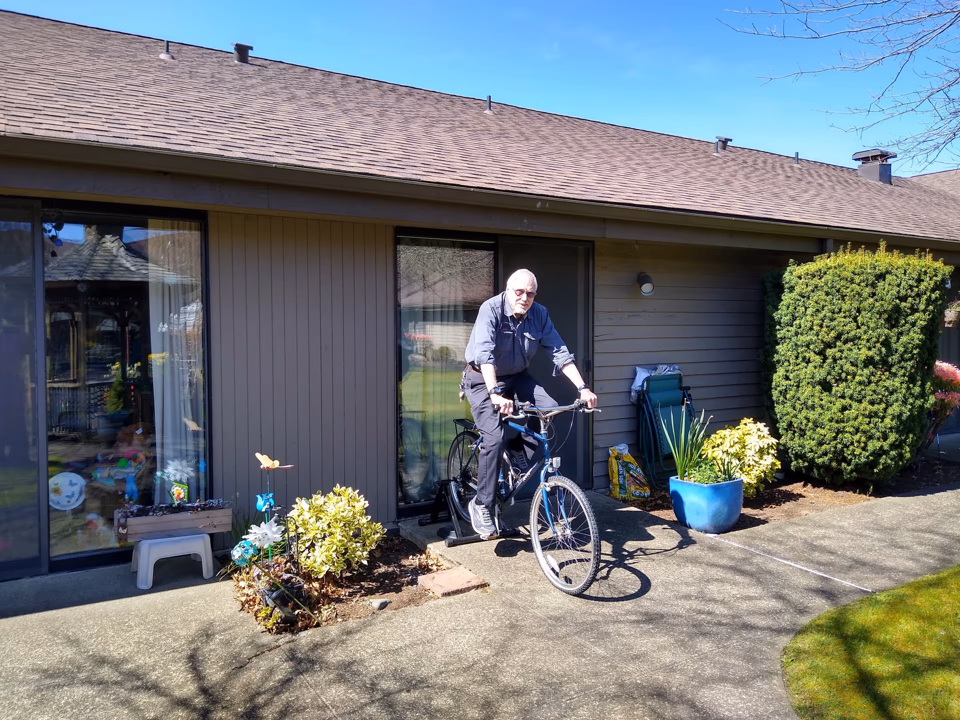 An elderly man with white hair and beard is riding a stationary exercise bike outside on a concrete pathway in front of a single-story building with sliding glass doors. There are potted plants, garden decorations, and shrubs around the area, and the sky is clear and blue.