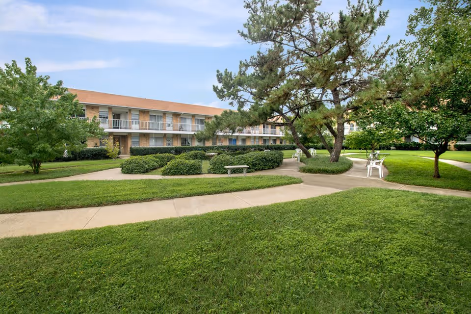 Outdoor view of Forest Dale Apartments showing a well-maintained green lawn with paved walkways, bushes, trees, and white outdoor chairs and tables. The apartment building with two floors and balconies is visible in the background under a partly cloudy sky.