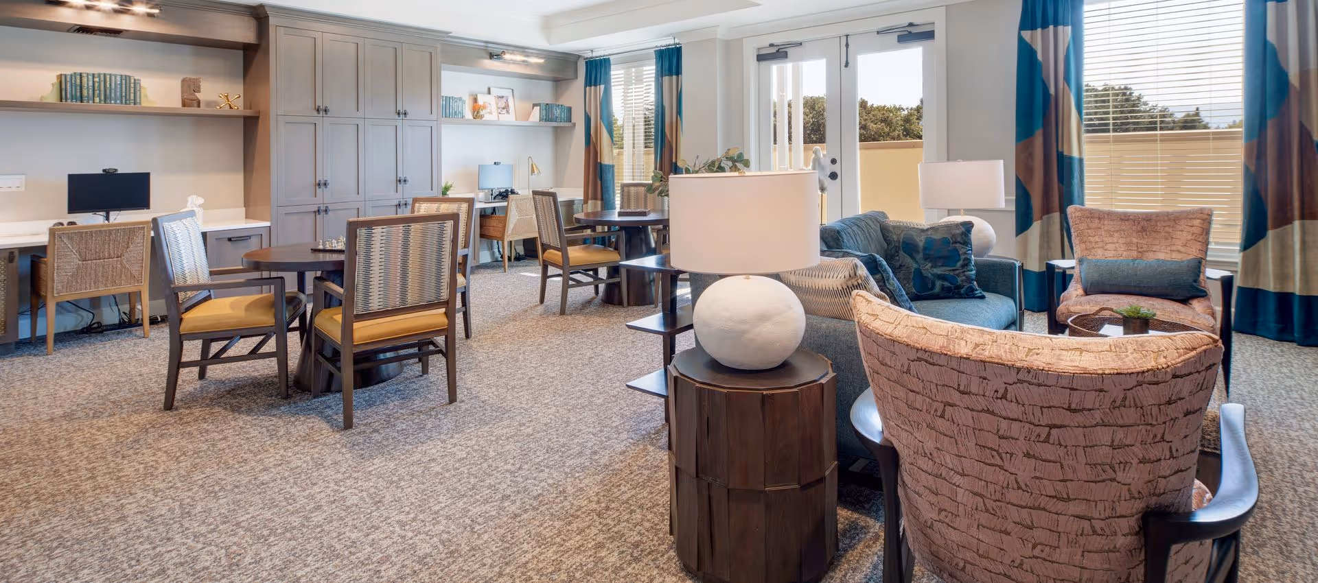 Bright communal lounge with sofas, armchairs, tables, desks and lamps arranged near French doors and windows.