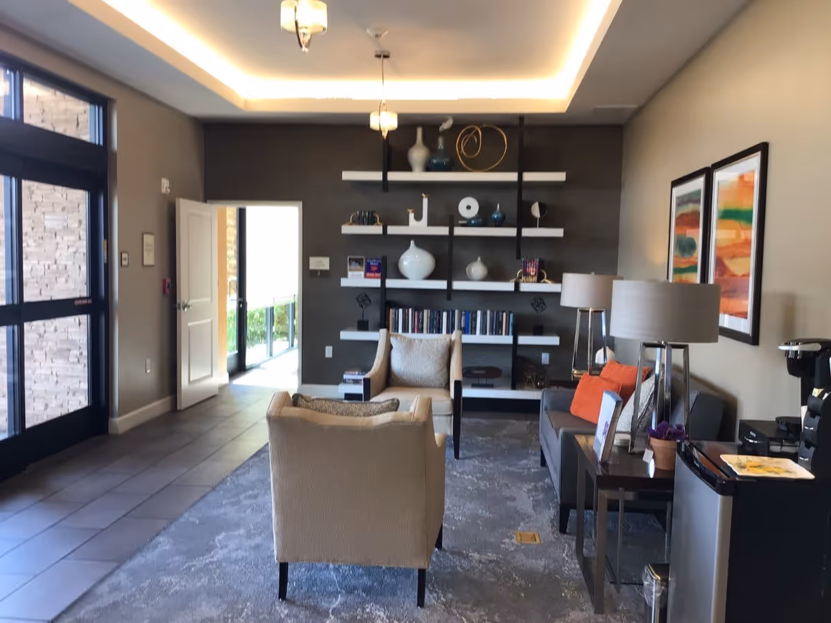 A cozy living room area in a senior living facility with two beige armchairs and a gray sofa with orange cushions. The room features a modern bookshelf with decorative items and books, two table lamps on side tables, framed abstract artwork on the wall, and a coffee station with a coffee maker and supplies. Large windows and a door provide natural light.