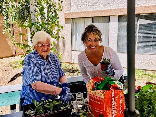 An elderly woman and a younger woman are gardening together outdoors at a senior living facility. They are smiling and wearing gardening gloves, surrounded by plants and gardening supplies including a bag of cactus, palm, and fern soil. The background shows a building with windows and some greenery.
