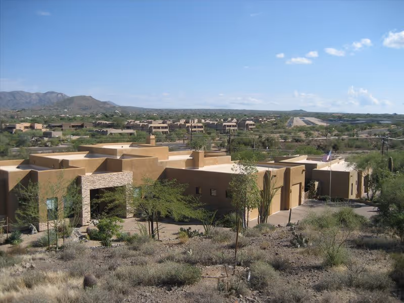 Exterior view of a southwestern-style assisted living facility with flat roofs and earth-toned walls, surrounded by desert vegetation and distant mountains under a clear blue sky.