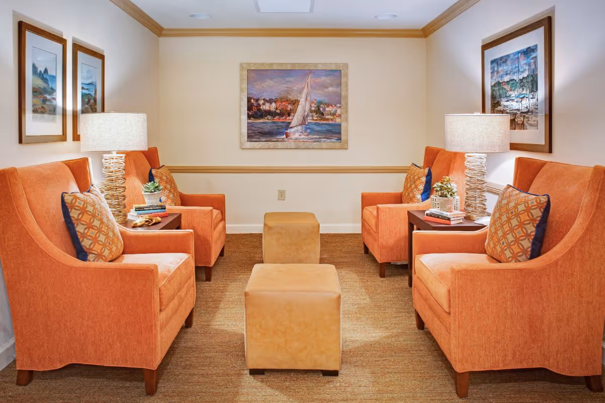 Small seating room with four orange armchairs, two ottomans, side tables with lamps, and framed artwork on the walls.
