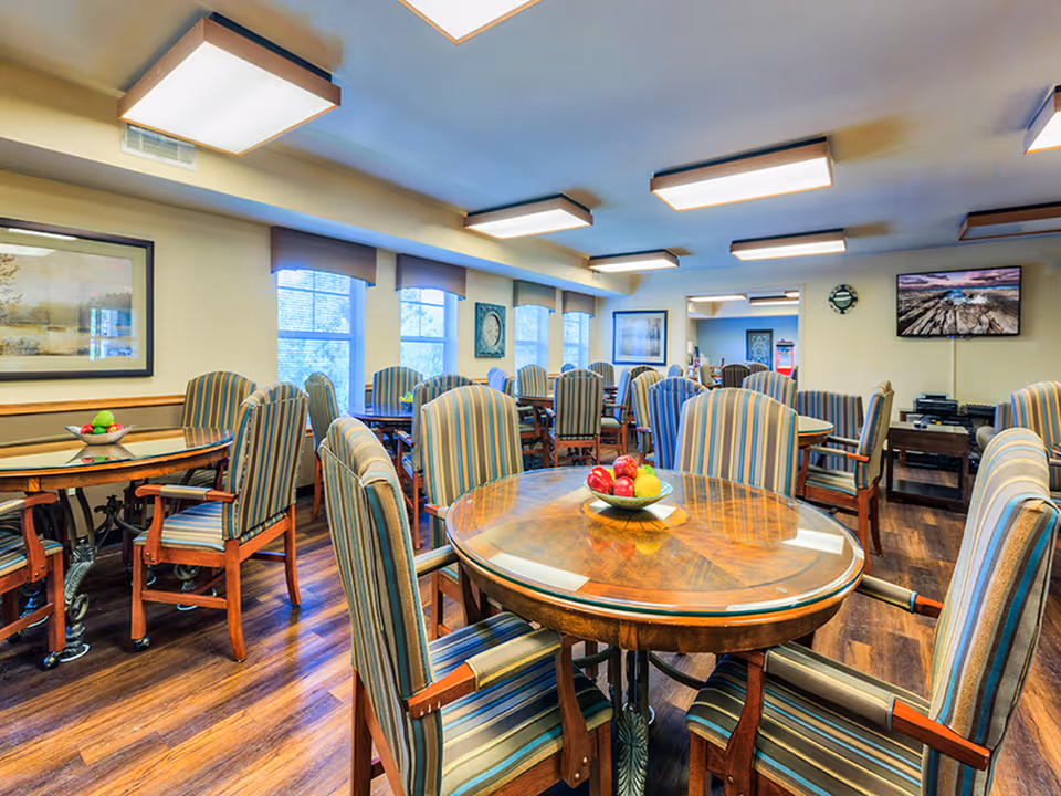 A well-lit dining room with multiple round wooden tables surrounded by striped upholstered chairs. Bowls of fruit are placed on the tables. The room has large windows with valances, framed artwork on the walls, a wall clock, and a flat-screen TV mounted on the far wall. The floor is wooden and the ceiling has rectangular light fixtures.
