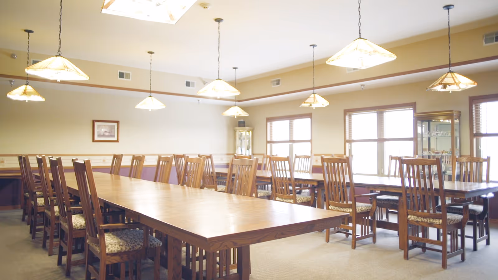 Large dining room with long wooden tables, multiple wooden chairs, and pendant lights over the tables.