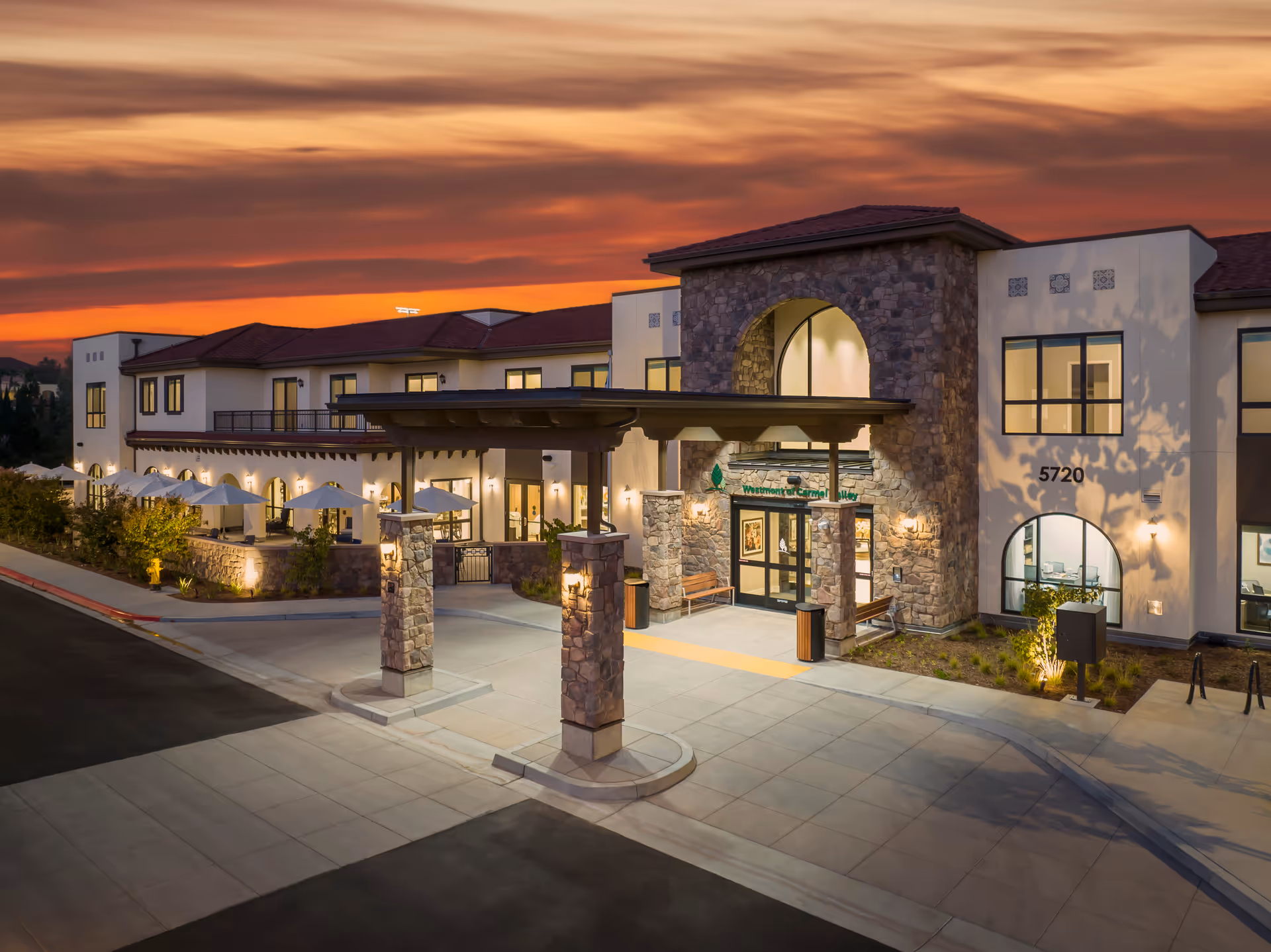 Exterior view of Westmont of Carmel Valley facility at sunset, showing a modern building with stone and stucco facade, large windows, a covered entrance with stone pillars, outdoor seating area with umbrellas, and well-lit landscaping.