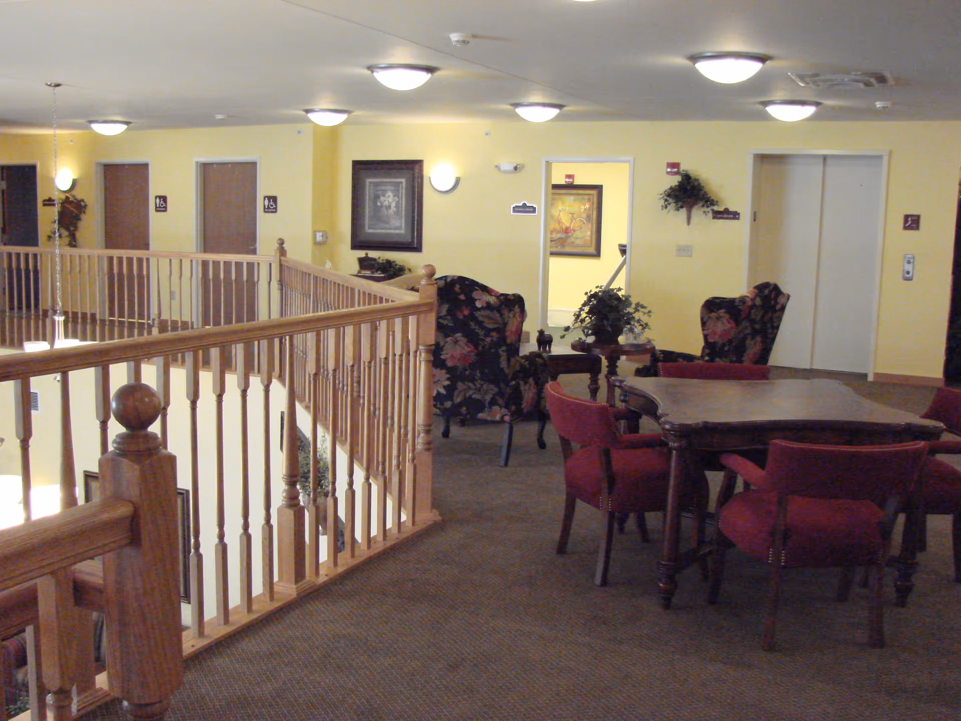 Upstairs open lounge with a wooden balustrade, a table surrounded by red chairs, floral armchairs, and an elevator and doors in the background.