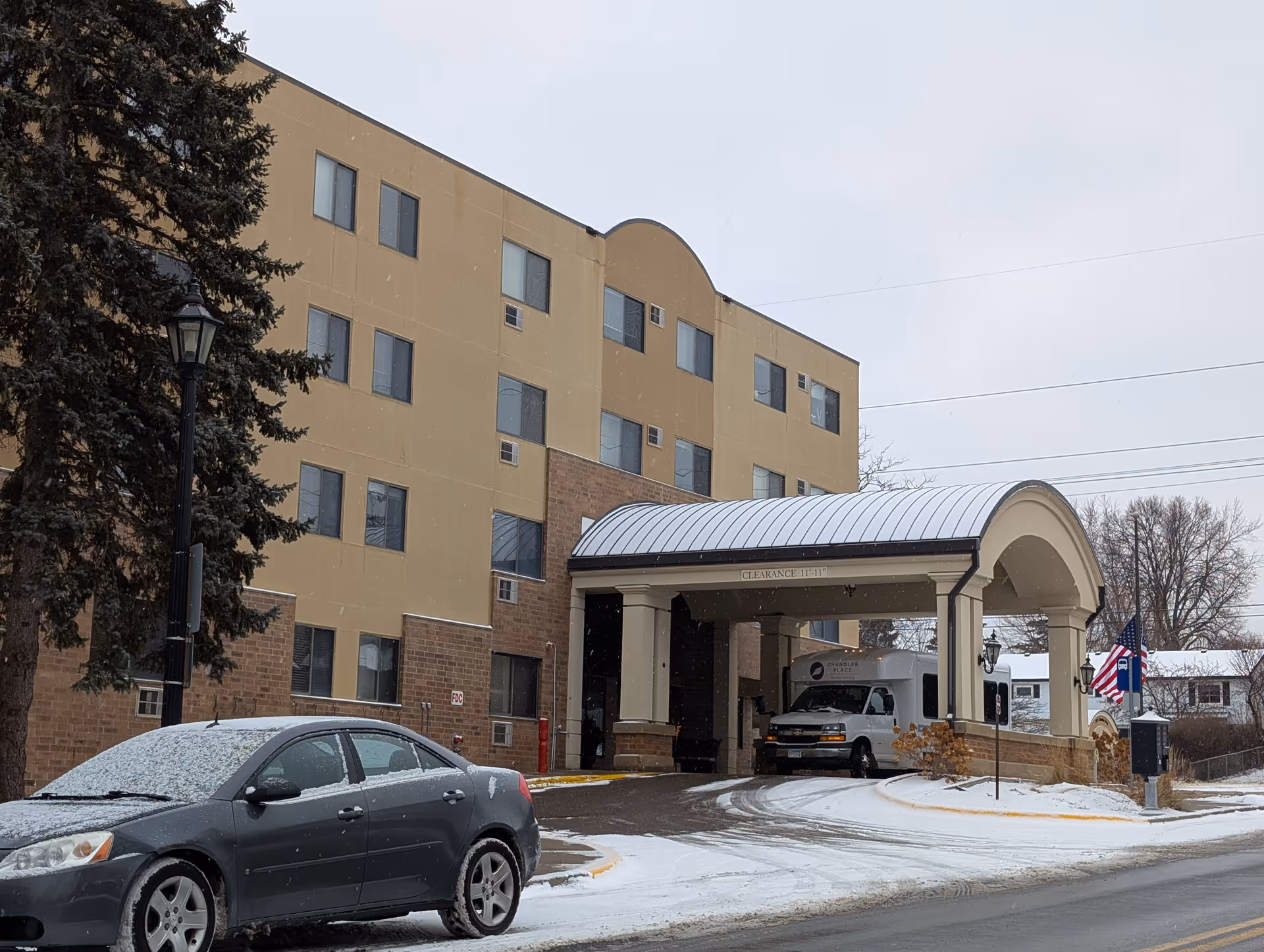 Exterior view of a multi-story senior living facility named Chandler Place on a snowy day. A covered entrance with a clearance sign is visible, along with a parked car and a van near the entrance. There is a large tree and an American flag near the driveway.