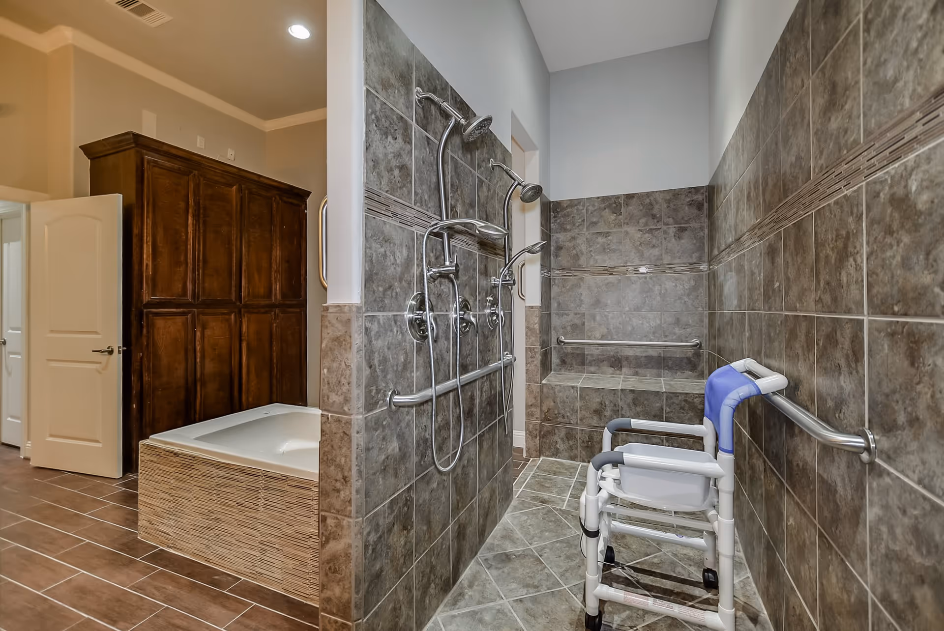 A spacious bathroom featuring a tiled walk-in shower with two showerheads and grab bars, a built-in tiled bench, and a white shower chair with wheels. Adjacent to the shower is a bathtub with a tiled surround and a large wooden cabinet. The floor is tiled in a brown and beige pattern, and the walls are painted light gray.