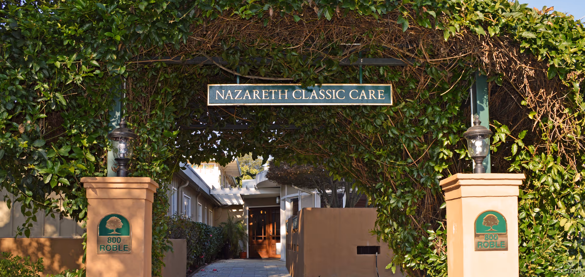 Entrance archway covered with green vines and leaves, with a sign reading 'NAZARETH CLASSIC CARE' hanging overhead. Two beige pillars on either side of the entrance display green plaques with a tree logo and the address '800 ROBLE'. A pathway leads to a building with a wooden door in the background.