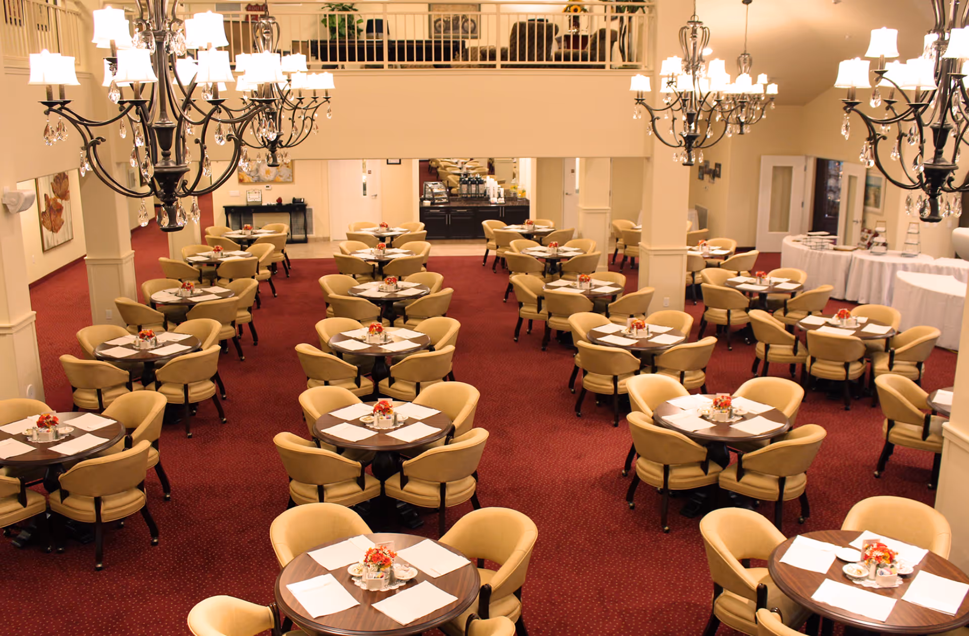 Large dining room with rows of round tables and beige chairs, chandeliers overhead, and a red carpeted floor.