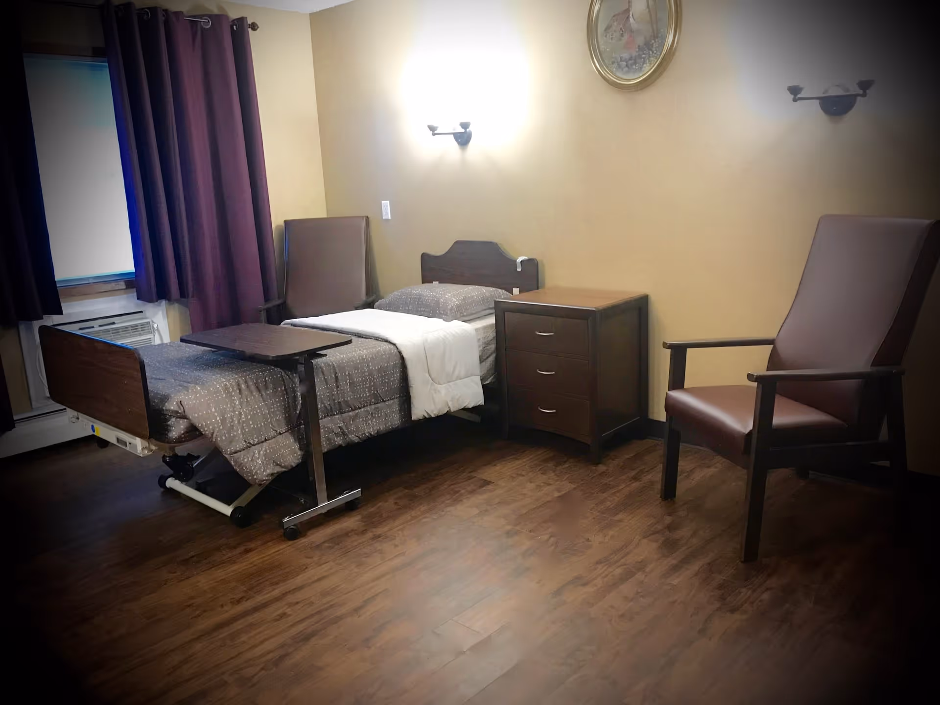 A senior living facility bedroom with a single hospital-style bed covered with a gray patterned blanket and white sheet. There is a small wooden bedside table with three drawers next to the bed. Two brown armchairs are positioned in the room, one near the window with dark purple curtains and the other near the wall. The room has wooden flooring and beige walls with two wall-mounted lights and a framed picture above the bed.