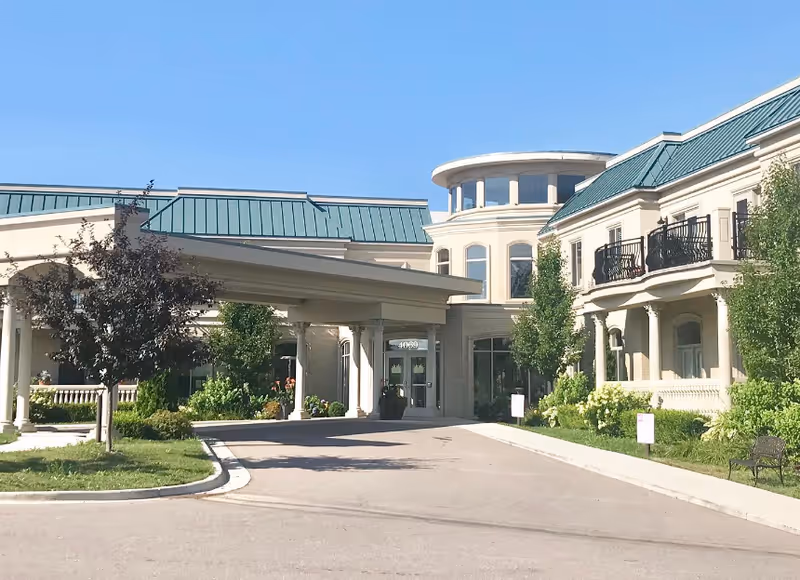Front exterior view of a senior living facility named Tranquility Estates with a covered entrance, green metal roof, beige walls, and landscaped greenery including trees and bushes under a clear blue sky.