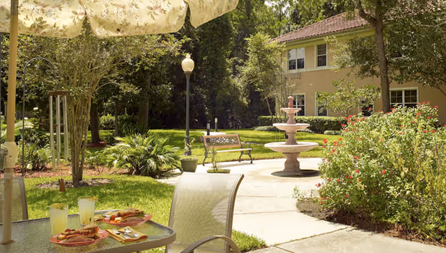 Sunlit courtyard with an umbrella-shaded patio table set for a meal, a central fountain, benches, and landscaped grounds beside a two-story building.