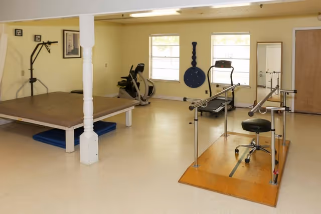 A rehabilitation room with exercise equipment including a treadmill, stationary bike, parallel bars with a stool, a padded platform, and a wall-mounted therapy device. The room has beige walls, two windows, and a wooden door.