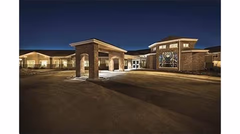 Nighttime exterior view of a brick building with a covered entrance and illuminated windows, featuring a large decorative circular window on the right side.