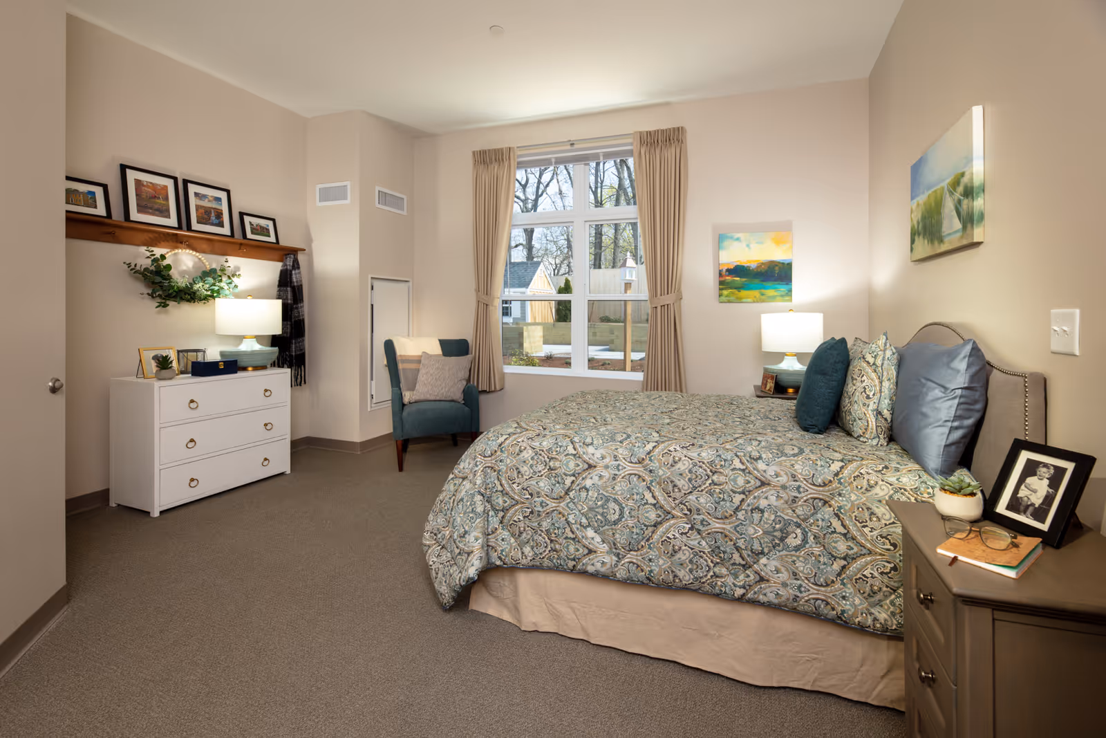 A cozy bedroom in a senior living facility featuring a neatly made bed with patterned bedding and multiple pillows. The room has beige walls and carpeted floors. There is a window with beige curtains letting in natural light. A teal armchair with a cushion sits near the window. On one side of the bed is a nightstand with a lamp, a framed photo, a small plant, and a book. On the opposite wall, there is a white dresser with a lamp, decorative items, and framed pictures on a wooden shelf above it. The room is warmly lit and decorated with colorful paintings on the walls.