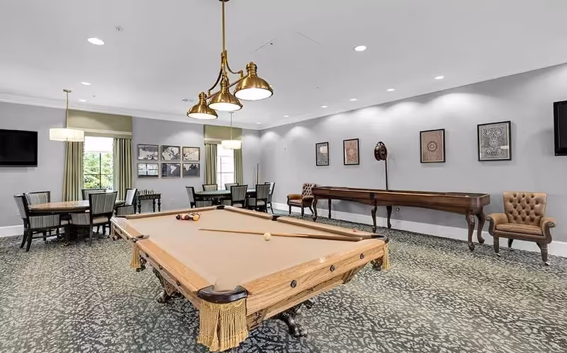 A bright interior recreation room with a pool table in the center, seating and card tables, a shuffleboard table, and framed wall art.