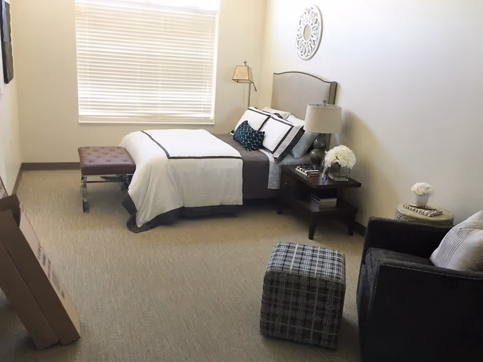 A neatly furnished bedroom with a bed, nightstand, armchair and window blinds.