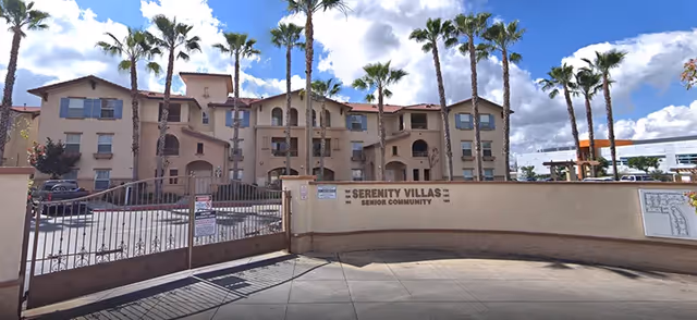 Exterior view of Serenity Villas Senior Community showing a gated entrance with a beige wall and multiple palm trees in front of a three-story building under a partly cloudy sky.