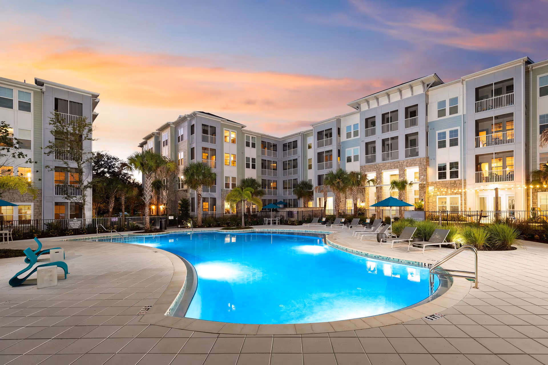 Outdoor swimming pool area at sunset with lounge chairs, umbrellas, and a multi-story residential building in the background with lit windows and palm trees around the pool.