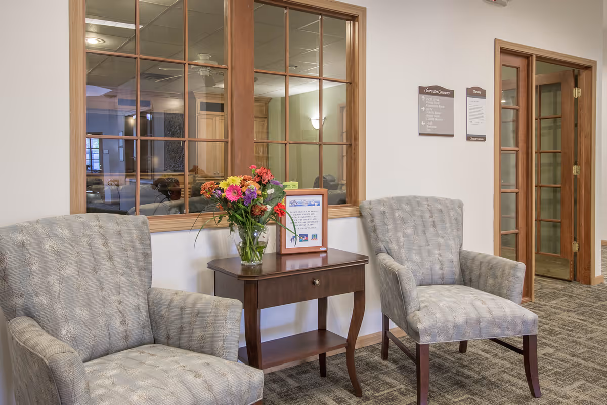 Two upholstered armchairs flank a small wooden table with a vase of flowers in a senior living facility seating area.