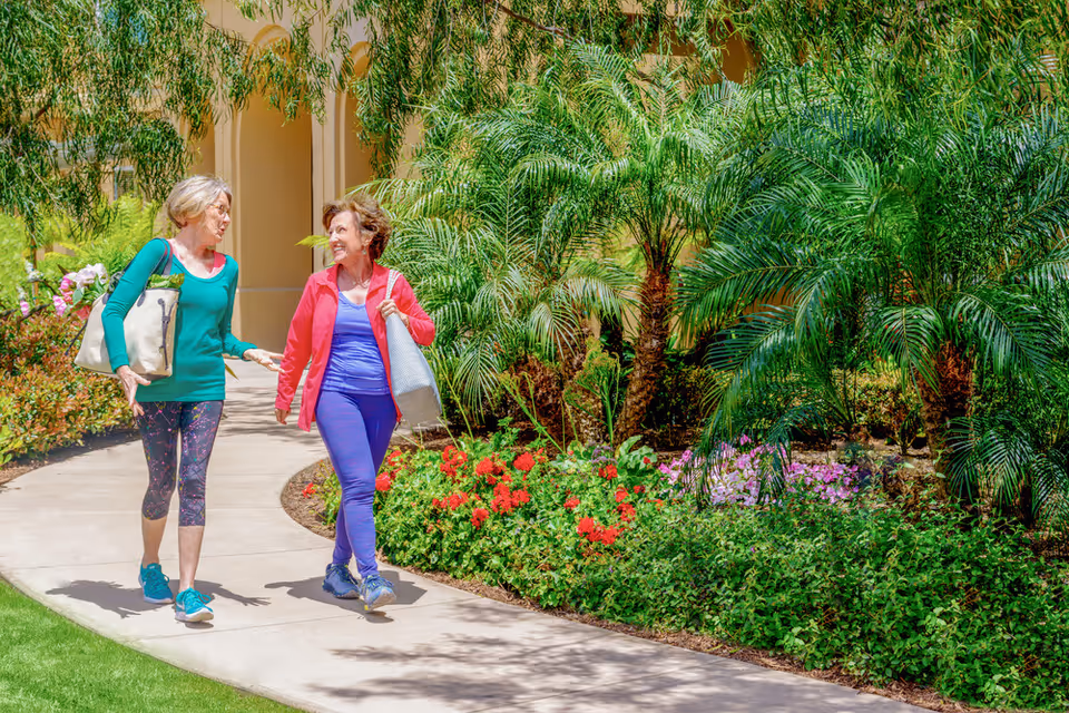 Two elderly women walking and talking on a curved garden pathway surrounded by lush greenery, palm plants, and colorful flowers at a senior living facility.