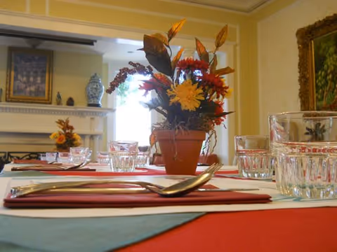 Close-up view of a dining table set with red placemats, silverware, and clear drinking glasses. A potted floral centerpiece with autumn-colored flowers is in the middle of the table. In the background, there is a fireplace with decorative items on the mantel and framed artwork on the walls.