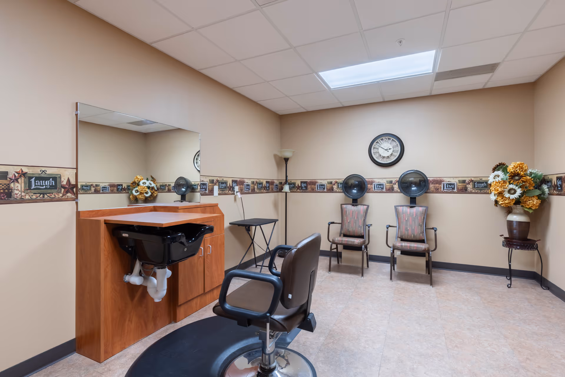 Interior view of a senior living facility hair salon area with a salon chair in front of a wash basin and mirror, two hair dryer chairs against the wall, a clock, a floor lamp, and a vase with artificial flowers.