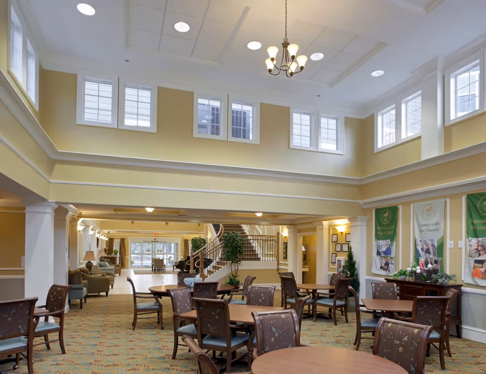 Spacious and well-lit common area in a senior living facility with multiple round tables and chairs arranged for socializing or dining. The room features high ceilings with multiple windows near the ceiling allowing natural light, a chandelier, and a staircase in the background. There are banners on the wall and comfortable seating areas along the side.
