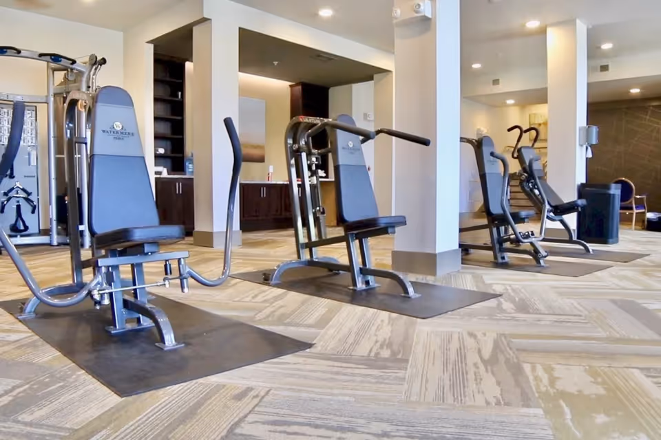 A fitness room with multiple exercise machines including seated weight machines, arranged on a patterned carpet floor. The room has white walls, pillars, and recessed lighting in the ceiling. Cabinets and shelves are visible in the background.