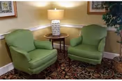 Two green upholstered armchairs flanking a round wooden table with a lamp in a cozy seating nook with framed art and a patterned rug.
