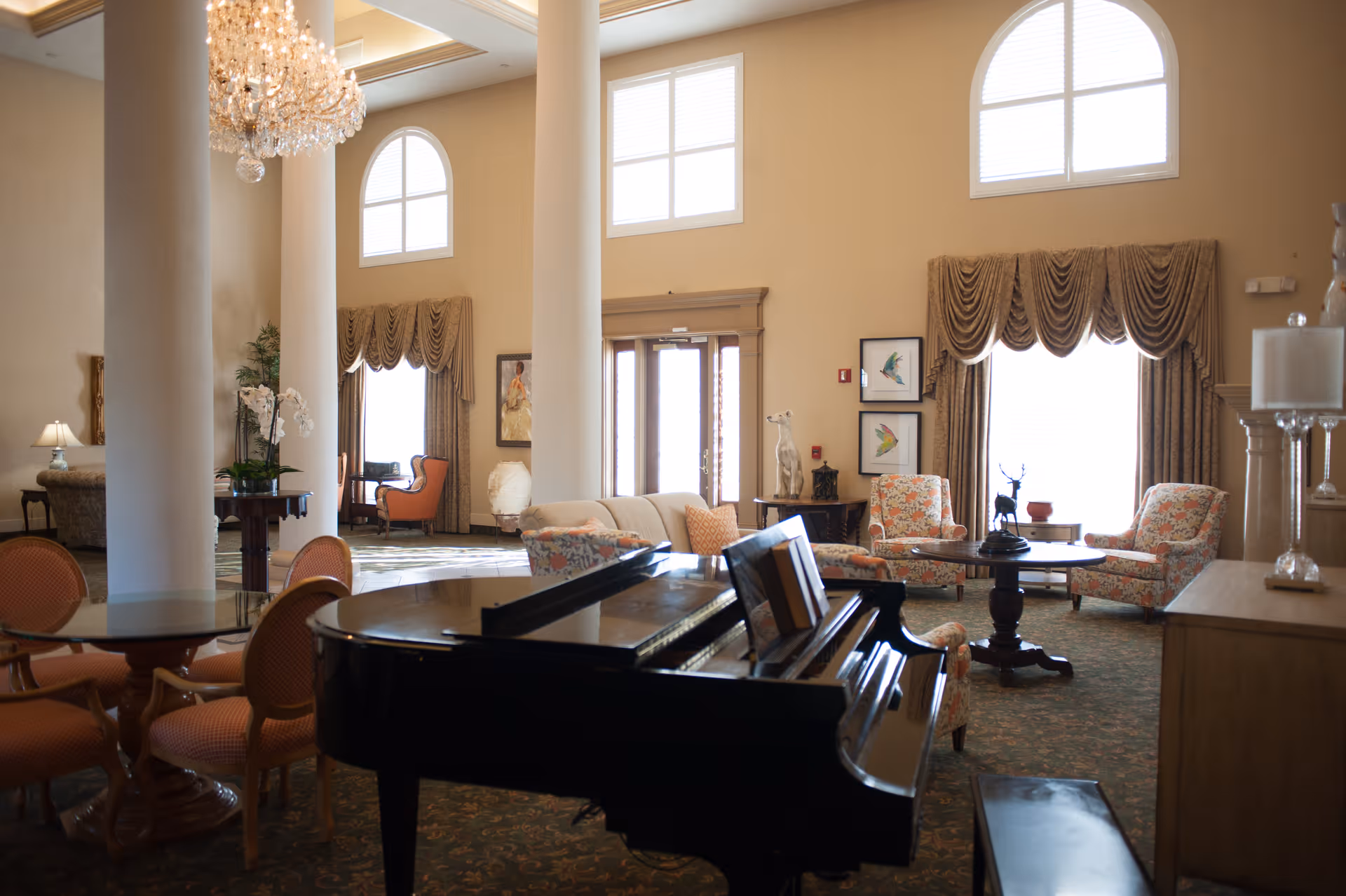 A spacious and elegant living room area with high ceilings, large arched windows, and a crystal chandelier. The room features a black grand piano in the foreground, several upholstered chairs with floral patterns, a round wooden table, and a glass-top table with four chairs. The walls are painted beige, and there are decorative curtains on the windows along with framed artwork on the walls.