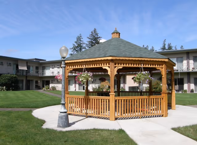 A wooden gazebo with hanging flower baskets sits in a grassy courtyard surrounded by a two-story retirement building.