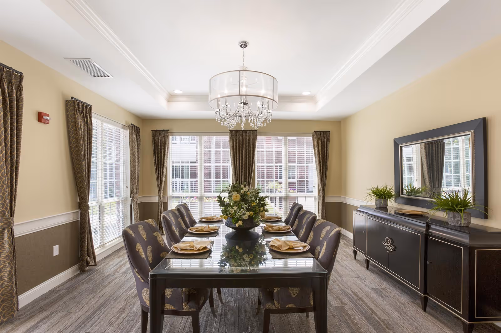 A well-lit dining room with a long glass table set for eight people, featuring patterned chairs and a floral centerpiece. The room has large windows with brown curtains, a chandelier hanging from the ceiling, and a black sideboard with a large mirror and two potted plants.