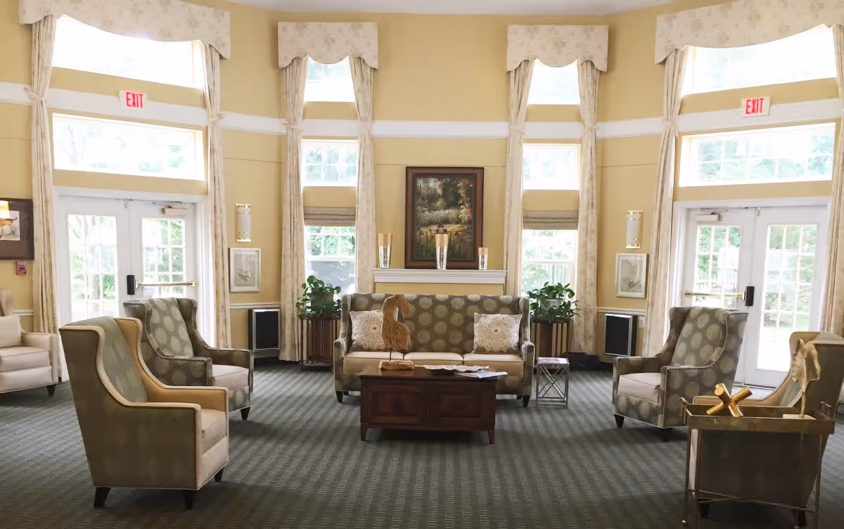 A spacious and well-lit senior living community lounge with large windows and glass doors letting in natural light. The room features a patterned carpet, a sofa with two decorative pillows, four armchairs arranged around a wooden coffee table with a decorative bird sculpture, and plants on either side of the sofa. The walls are painted a soft yellow with white trim, and there are framed pictures and sconces on the walls. Exit signs are visible above the doors.