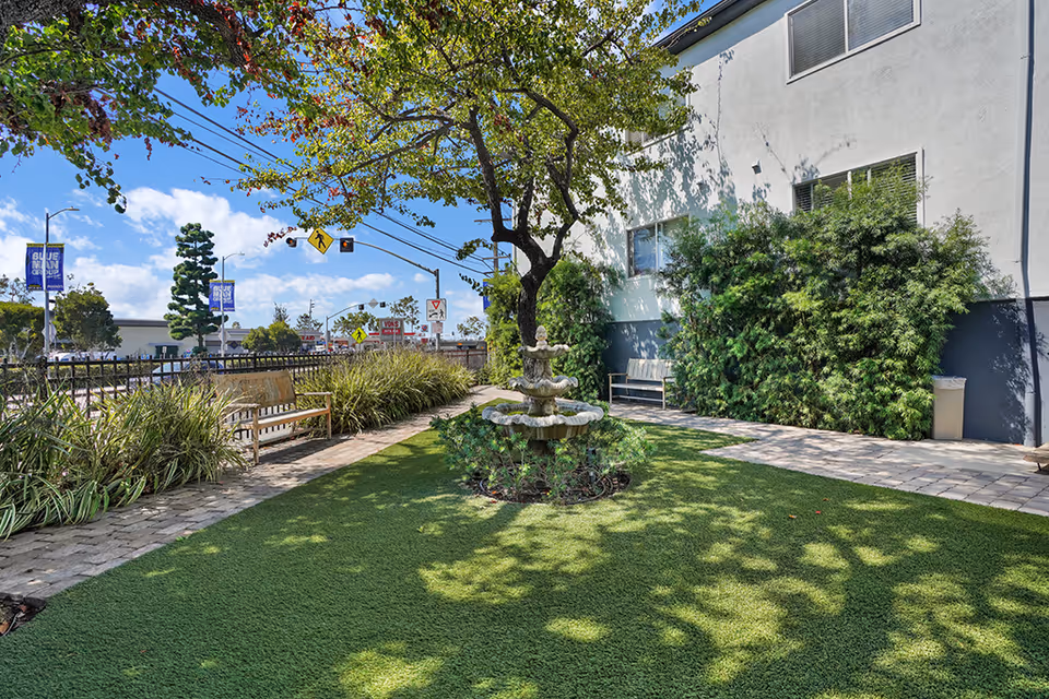Outdoor garden area at Terraza of Cheviot Hills featuring a green lawn with a decorative tiered fountain in the center, surrounded by trees, bushes, and benches along a paved walkway. The building exterior is visible on the right side, and a street with traffic signs and power lines is seen in the background under a blue sky with scattered clouds.