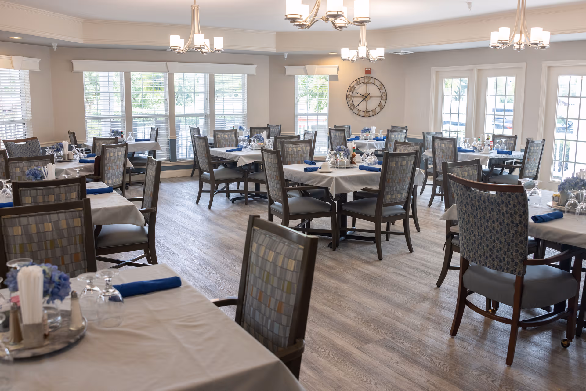 Spacious dining room with multiple tables set with white tablecloths, blue napkins and place settings, surrounded by chairs and large windows letting in daylight.