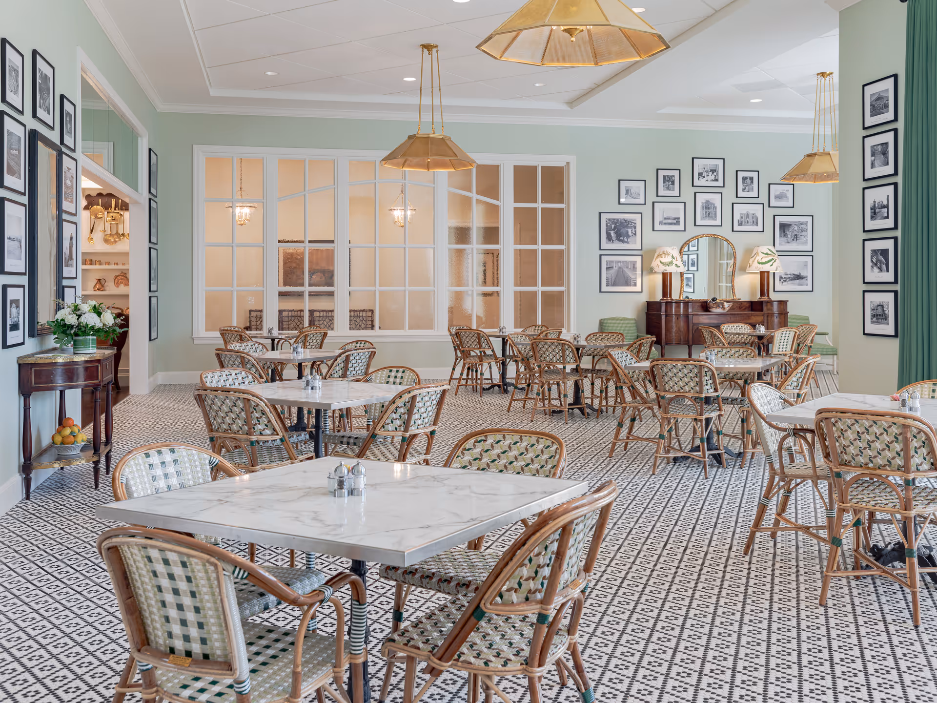 A bright and elegant dining room with multiple square marble-top tables surrounded by woven rattan chairs. The room features a patterned tile floor, light green walls adorned with numerous black-and-white framed photographs, and two gold pendant lights hanging from the ceiling. There is a wooden sideboard with two lamps and a mirror, and a small table with a vase of flowers and a bowl of fruit near the entrance.