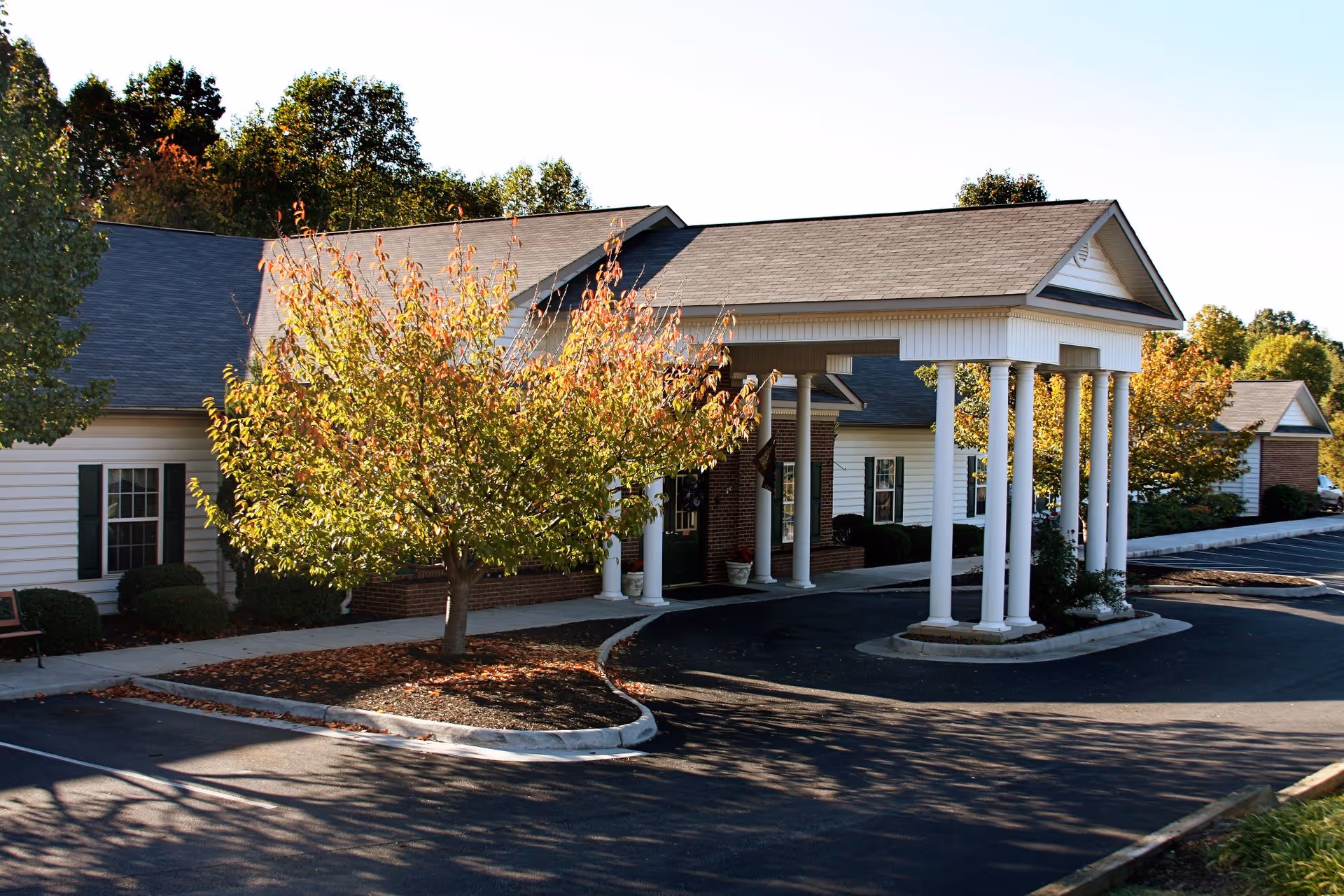 Exterior view of Heritage Green Assisted Living and Memory Care facility showing a covered entrance with white columns, a tree with autumn foliage, and a parking area.